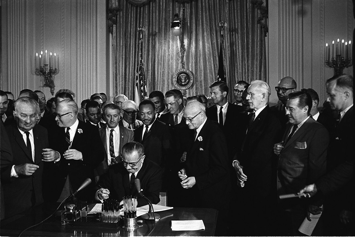 President Lyndon B. Johnson signs the 1964 Civil Rights Act as Martin Luther King, Jr., and others, look on. 