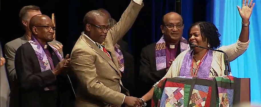 The Rev. Cynthia Moore-Koikoi held the hand of her husband– the Rev. Rafael Koikoi — as she accepted her election as bishop by the Northeastern Jurisdiction. Her bishop, Baltimore-Washington Episcopal Area Bishop Marcus Matthews, left, applauds. Boston Episcopal Area Bishop Sudarshana Devadhar stands behind her. Photo courtesy of Baltimore-Washington Conference. 
 

