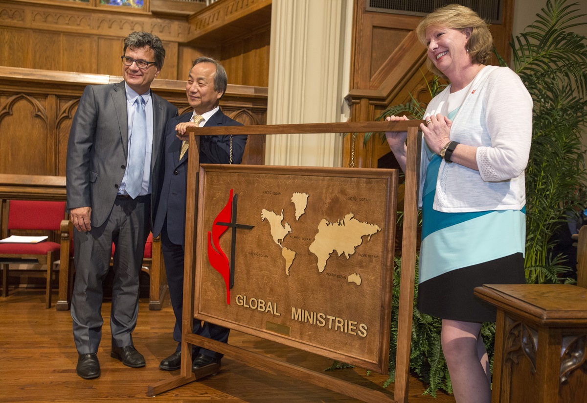 Bishop Sue Haupert-Johnson from the North Georgia Conference presents a wooden map featuring continents and the United Methodist cross and flame to Thomas Kemper and Bishop Hee-Soo Jung following the dedication inside Grace United Methodist Church. 
