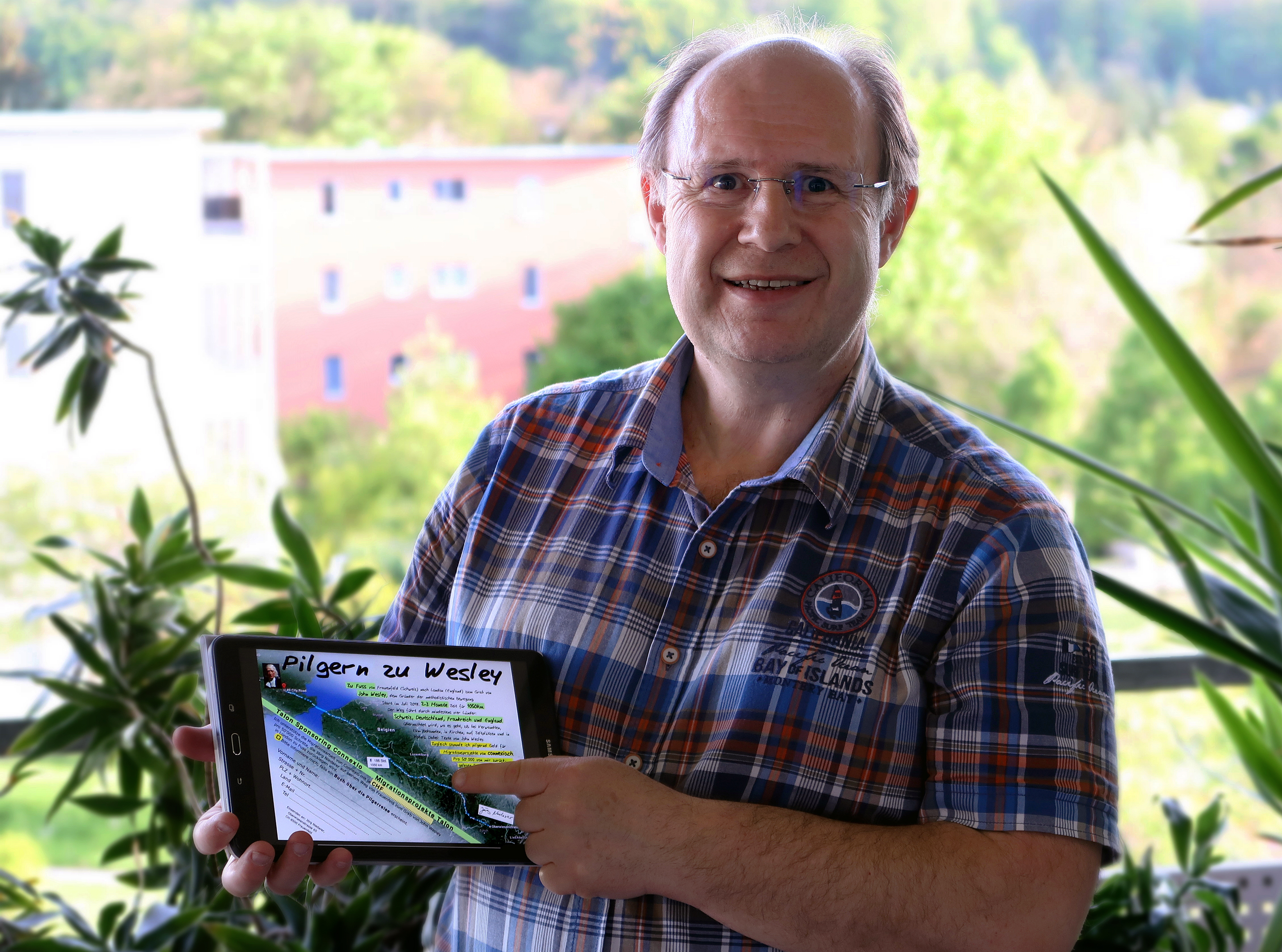 Jörg Niederer stands on the balcony at his home in Frauenfeld, Switzerland, holding a tablet that shows the route he will take on his pilgrimage across Europe to John Wesley’s gravesite in London, England. Photo courtesy of Jörg Niederer.