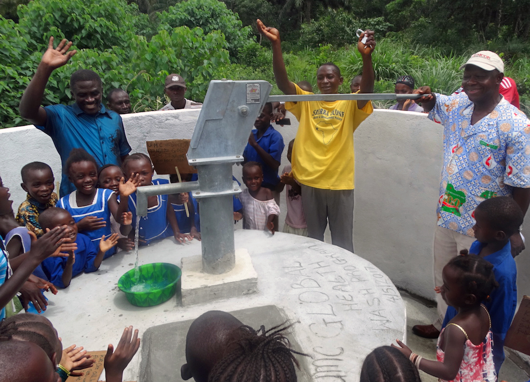 Children and families clap in celebration as Bishop John K. Yambasu (far right in light blue shirt) pumps the first water from a new well during a dedication ceremony in Manjama in the Bo District of Sierra Leone. A Global Health agency initiative provided each community a four-seat toilet and a hand-dug well for United Methodist primary schools in Manjama and Fulawahun. Photo by Phileas Jusu, UMNS.
