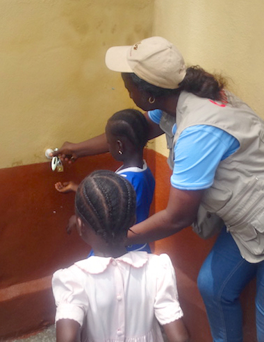 Health Coordinator Catherine Norman (center in cap) shows children of the United Methodist Church Primary School in Fulawahun, Sierra Leone, how to use a water faucet to wash their hands after using the toilet, while Bishop Yambasu (far right) looks on. A United Methodist Global Health agency initiative has provided much needed toilets and wells for primary schools in two rural communities that previously had a prevalence of diarrhea and worm infestations due mainly to lack of toilets and good drinking water. Photo by Phileas Jusu, UMNS.