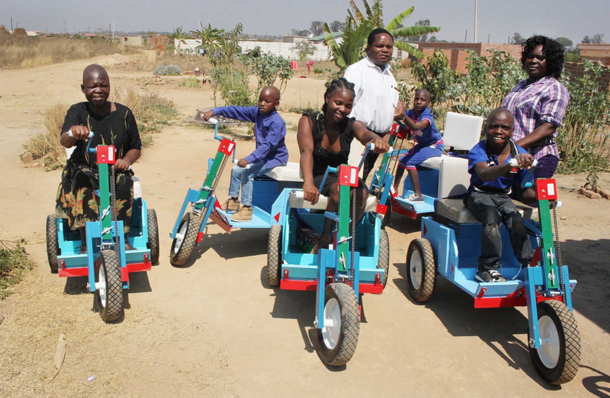 Disabled children test out five of the wheelchairs distributed by Raphael Bareyi (in background) in Stoneridge, Zimbabwe. The children are Tanaka Jokonia, Rodrick Ngwaru, Tsitsi Dzengu, Natasha Dziva, Kerry Musana. Standing to the right is Rachel Kaziti Madzana. Photo by Chenayi Kumuterera, UMNS.  