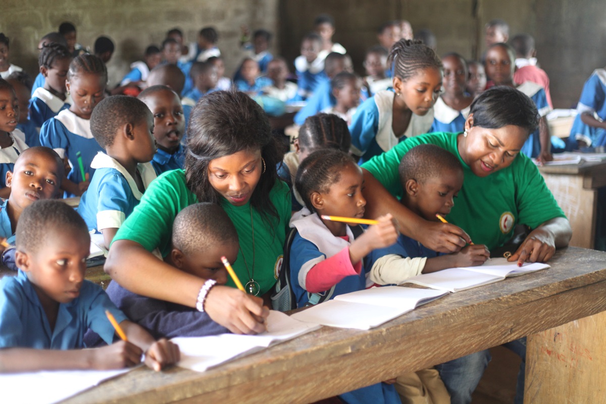 The Rev. Melvis Abah, a United Methodist pastor, and Collette Ndobe with the United Methodist Women Association in Cameroon show the children of Government English Primary school Monatele how to write on paper with their pencils. Photo by Marius Bonfeu.