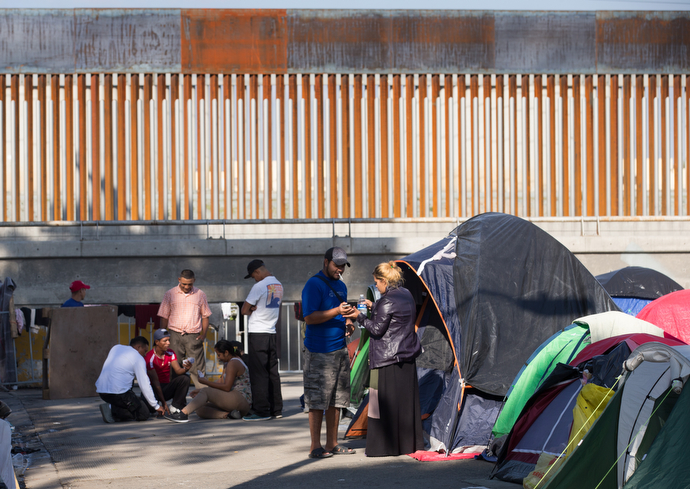 Migrants from Central America pass the time in a makeshift camp outside the Benito Juarez sports complex in Tijuana, Mexico. Although many people in the migrant caravan left this facility for another camp with better sanitary conditions, others remained here because the sports complex is only yards away from the U.S. border fence visible in the background.