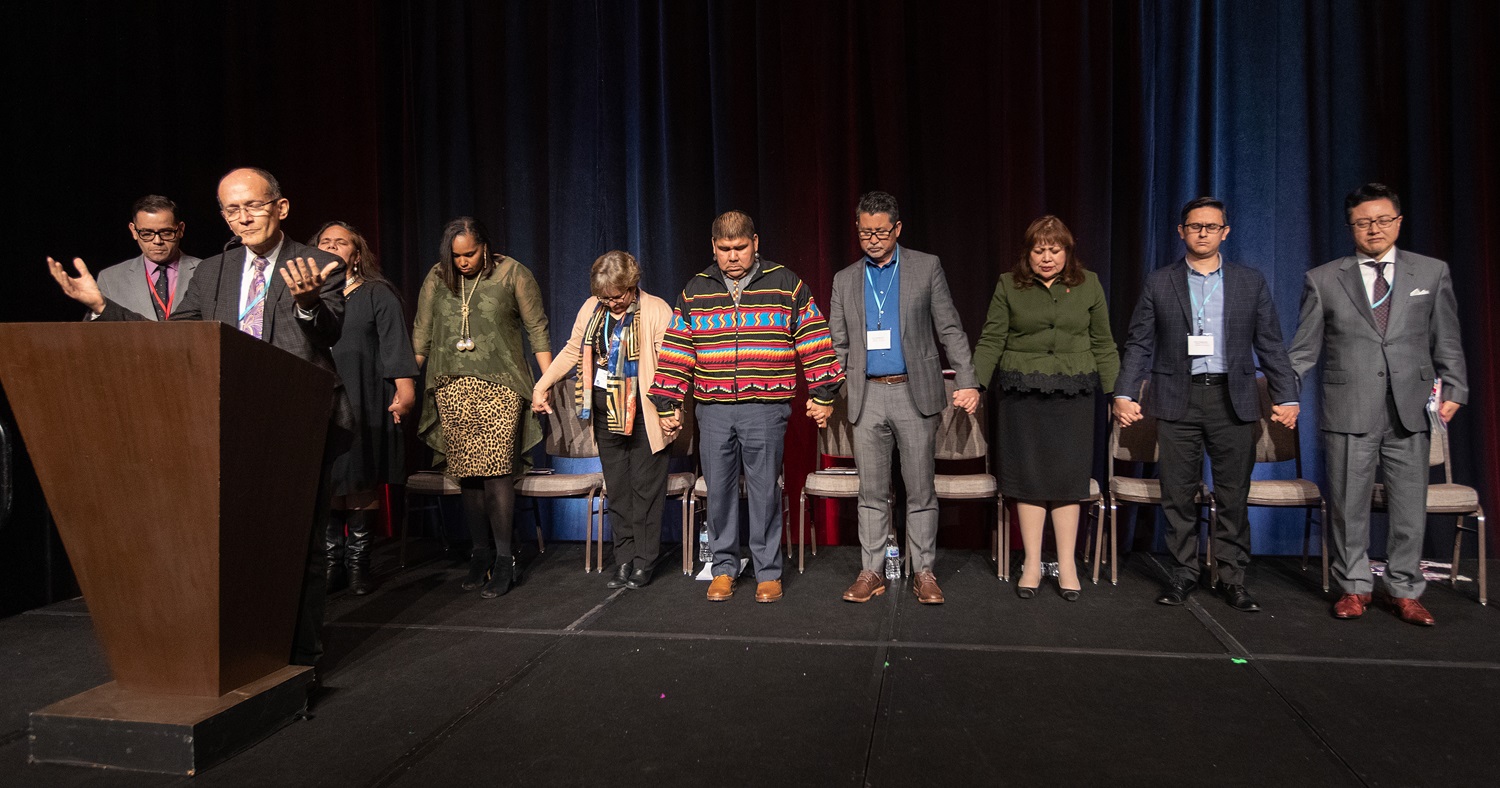 El Rev. Jorge Domingues (en el podio) dirige una oración por los representantes de las iniciativas étnicas de la Iglesia Metodista Unida durante la sesión informativa de la Conferencia Pre-General 2020 en Nashville, Tennessee. Domingues es director ejecutivo de ministerios conexos para la Conferencia California-Nevada. De derecha a izquierda: Rev. Paul Chang, Director Ejecutivo del Plan para el Ministerio Coreano; Manuel Padilla, Director Interino del Plan Nacional para el Ministerio Hispano-Latino; Obispa Minerva Carcaño, líder episcopal de la Conferencia Anual de California-Nevada; Rev. John Oda, Gerente del Plan para el Ministerio de Lengua Asiático-Americana; Rev. Chebon Kernell, Director Ejecutivo del Plan Ampliado Nativo-Americano; Revda. Lyssette Perez, Pastora en la IMU Oasis en Pleasantville, Nueva Jersey y presidenta de MARCHA; ; Toska Medlock Lee, Directora Interina del Plan de Fortalecimieno de la Iglesia Negra para el siglo 21; Monalisa Tuitahi, Directora Ejecutiva el Caucus Nacional de las Comunidades Isleñas del Pacífico y el Rev. Gustavo Vasquez es el Director de Noticias Metodistas Unidas para la audiencia Hispano-Latina.  Foto de Mike DuBose, Nptocoas MU.