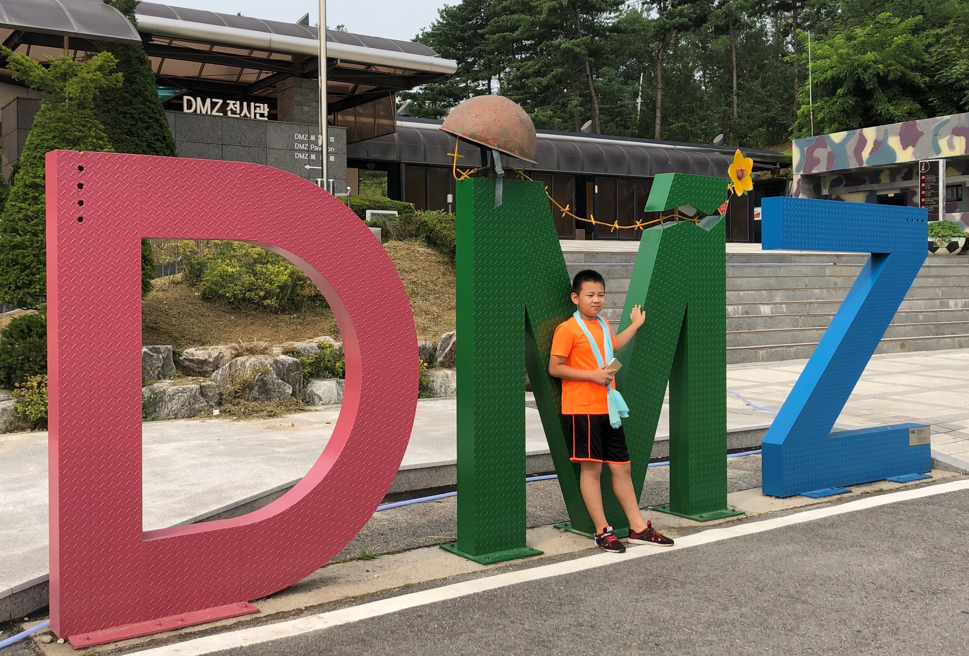 한 관광객 어린이가 DMZ (군사분계선, Military Demarcation Line) 전시관 앞에서 사진을 찍고 있다. 가운데 M자 위에 놓인 녹슨 철모와 꽃이 인상적이다. 사진, 김응선 목사, 연합감리교뉴스. 