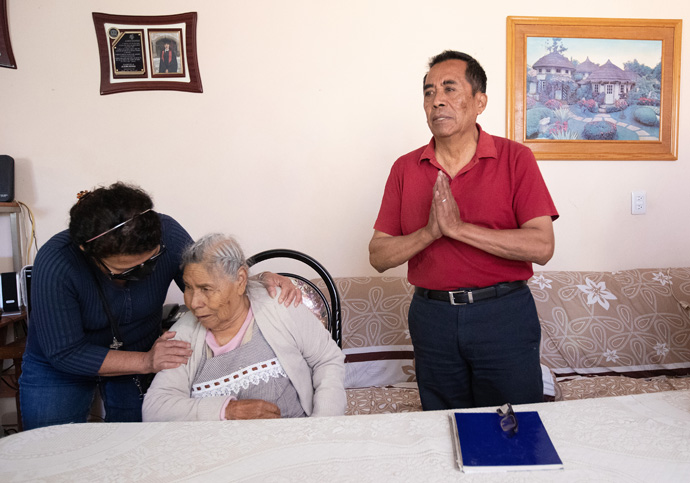 Pastor David Castro Fernández expresses his gratitude for the unexpected ministry he and his family developed by helping northbound migrants who were riding freight trains that stopped behind their home in Tlaltelulco, Mexico, and for a visit from leaders of the Methodist Church of Mexico and The United Methodist Church who came to hear their story. At left, the Rev. Toña Rios prays with Fernández’s mother-in-law, Heruelinda George Sanchez, 85, who spent many years helping care for the migrants. Photo by Mike DuBose, UM News.