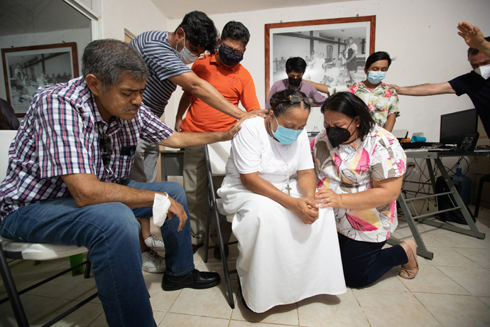 Leaders from the Methodist Church in Mexico pray with Olga Sánchez Martínez (center), founder of the Jesús el Buen Pastor migrant shelter in Tapachula, Mexico. Bishop Raquel Balbuena (kneeling) led the prayer. She was joined by (from left) Bishop Felipe Ruiz Aguilar and the Revs. Uzi Castañeda Morales and Isidro Martínez Cortés. At right is the Rev. Arturo González Sandoval, president of the National Commission on Migration Issues, praying with two staff members of the shelter. Photo by Mike DuBose, UM News.