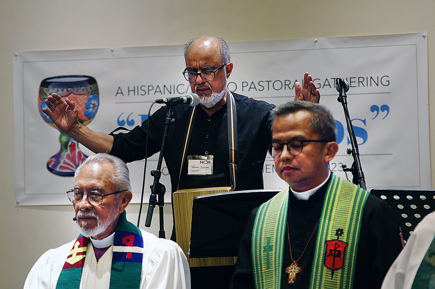 El Rev. Tercio Junker (centro), pastor en la Conferencia Anual del Norte de Illinois, dirige la oración para consagrar los elementos del sacramento de la comunión. Abajo a la izquierda el Obispo Elías Galván, actualmente retirado y fundador de la Conferencia Anual del Desierto del Suroeste que actualmente lidera el Obispo Carlo Rapanut a la derecha de la imagen.