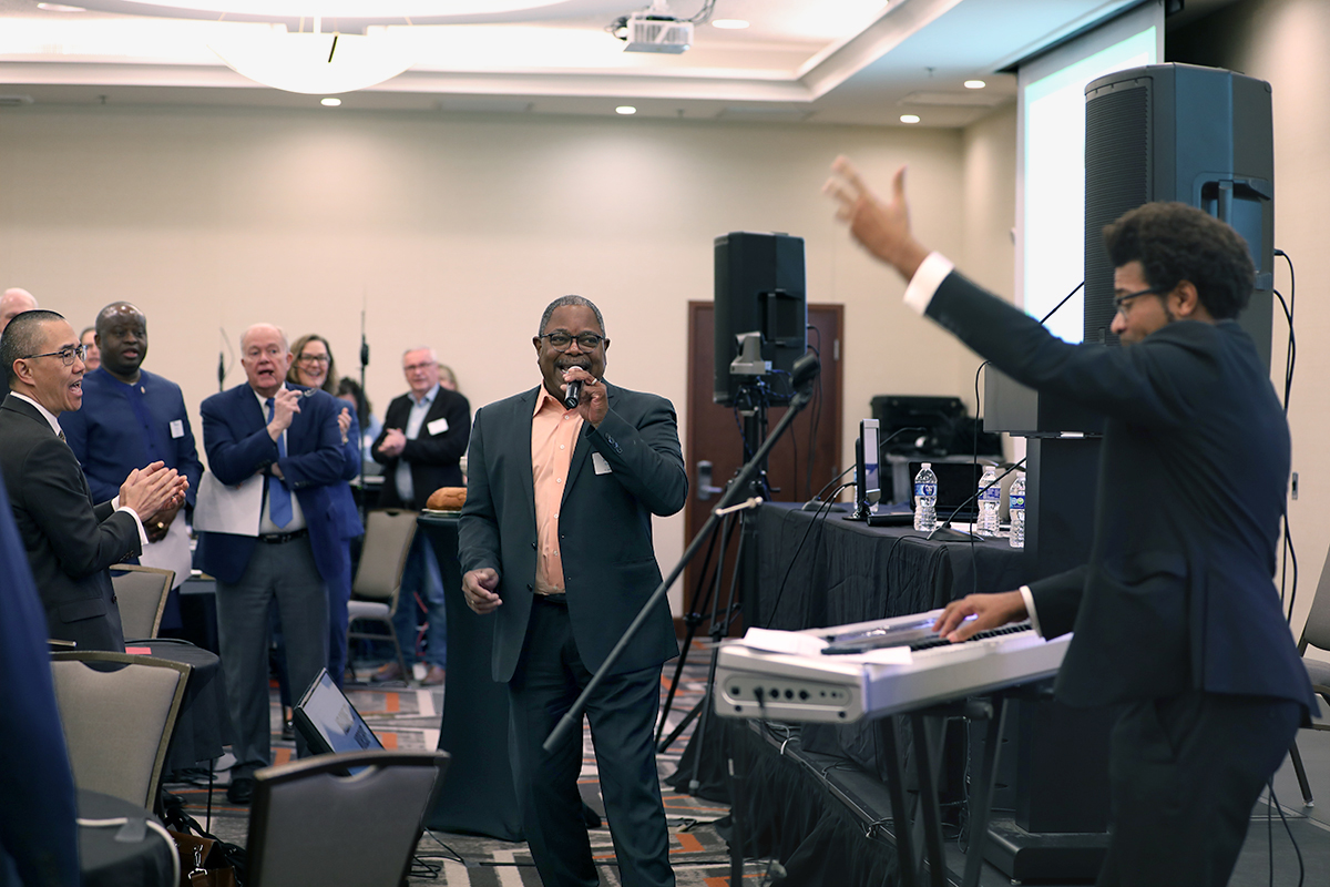 O Rev. Reggie Clemons, membro do Conselho Geral de Finanças e Administração (centro, com microfone), e o Rev. Theon Johnson III, membro da Mesa Conexional (no teclado), unem-se para liderar o culto numa reunião conjunta de 19 a 20 de Fevereiro - reunião para finalizar o orçamento denominacional proposto para a Conferência Geral.