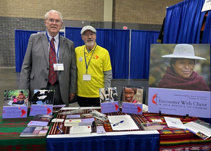 Lloyd Ambrosius (left) joins the Rev. Doug Ruffle at the Encounter with Christ in Latin America and the Caribbean booth in the exhibition hall at the General Conference in Charlotte, North Carolina. Ambrosius died May 7 at age 82. He had a key role in founding and growing Encounter with Christ in Latin America and the Caribbean, an endowment fund for Methodist ministry south of the U.S. Photo courtesy of the Rev. Cynthia Weems.