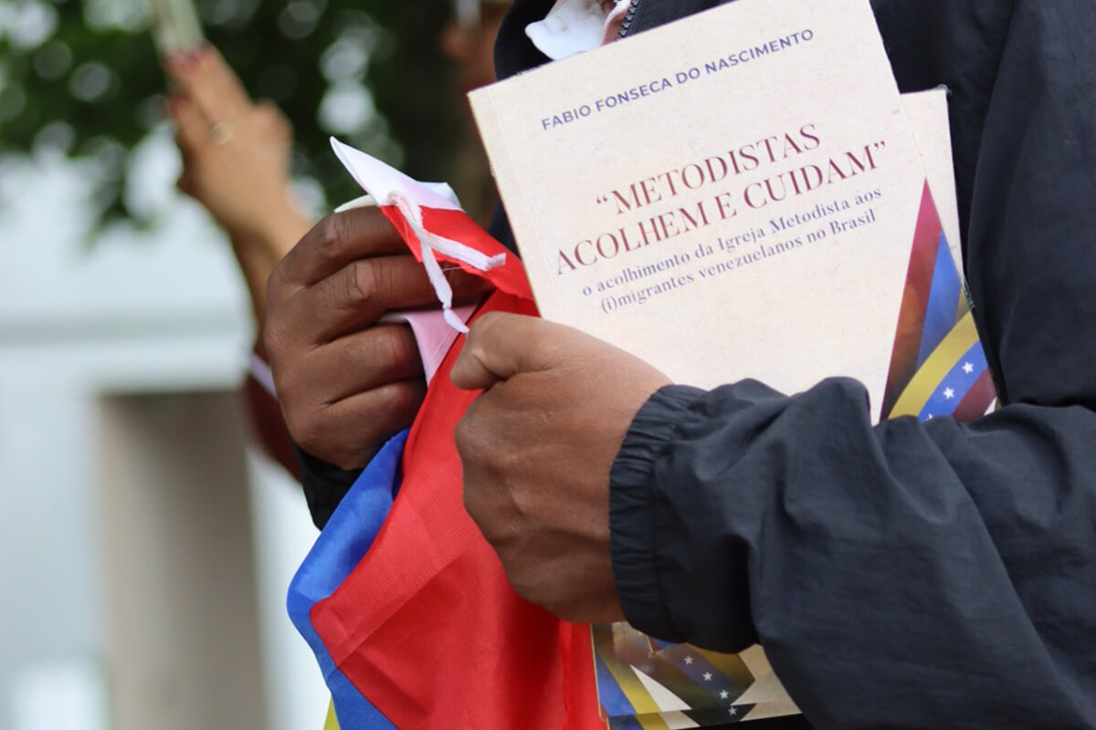 Durante una conversación y caminata con el autor, el Pastor Edson César da Silva porta el libro "Metodistas Acogen y Cuidan" y la bandera de Venezuela, país de donde proviene la mayor parte de las personas atendidas por el proyecto. Foto de Sara de Paula, para Noticias MU