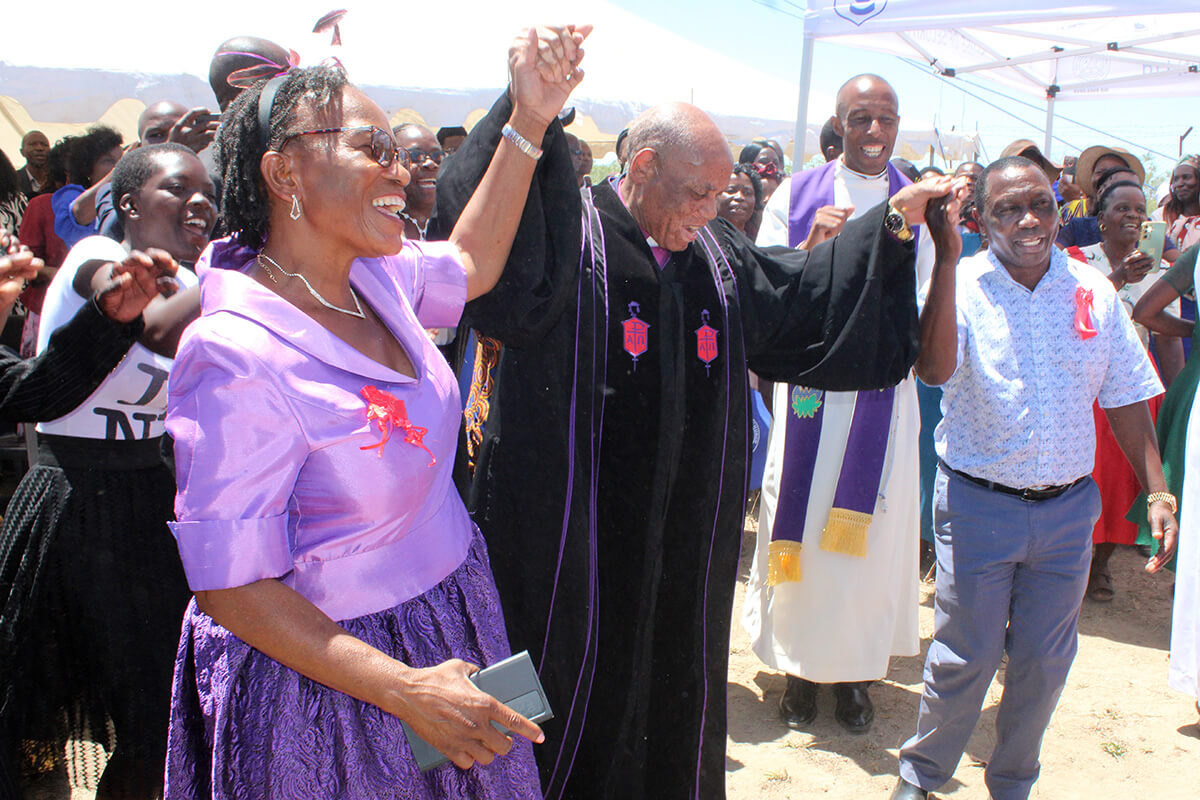 Patricia and Ray Kaukonde join hands with Bishop Eben K. Nhiwatiwa to celebrate the dedication of Donzwe United Methodist Church near Mudzi, Zimbabwe, in December 2024. Ray Kaukonde fulfilled his dying mother’s wish by completing construction of the church that was started in 1977 by his grandfather. Photo by Kudzai Chingwe, UM News.
