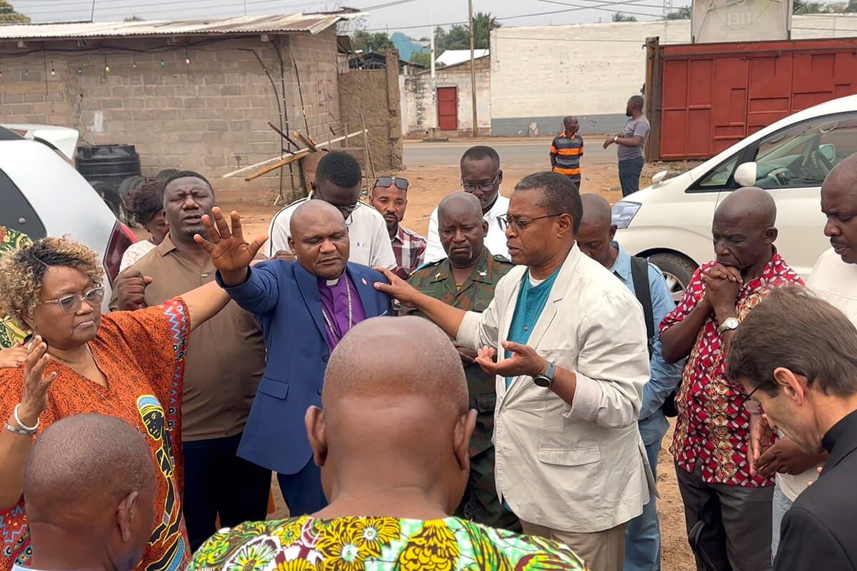 Bishop Nelson Kalombo Ngoy of the Tanganyika Episcopal Area (center, in blue suit) and the Rev. Alpher K. Sylvester of the New York Conference (right, in light jacket) stand amid a group of clergy and community members with hands raised in prayer during an outreach visit in Kalemie, Congo. Photo by Chadrack Tambwe Londe, UM News.