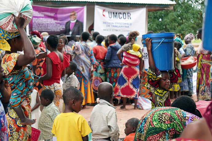 A young girl carries a bucket of supplies on her head amid a crowd of beneficiaries gathered in front of banners from UMCOR and the Tanganyika Episcopal Area. The distribution of relief kits aims to restore the dignity of families in Nyunzu, who are struggling to survive in the face of food insecurity and disease resulting from recent floods and conflicts in eastern DRC. Photo courtesy of the Disaster Management Office of the Tanganyika Episcopal Area.