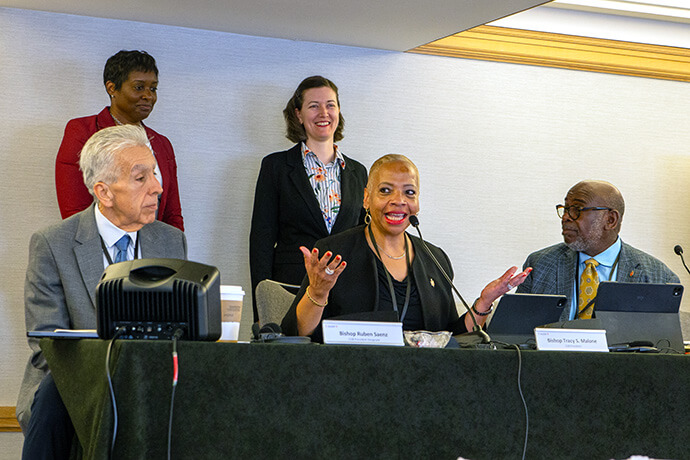 La révérende Aleze Fulbright (à l’arrière gauche), secrétaire sortante de la Conférence Générale, et Emily Allen, la nouvelle secrétaire par intérim, se tiennent derrière la présidente du Conseil des évêques, Tracy S. Malone, tandis que celle-ci remercie Fulbright pour le travail d’organisation de la Conférence Générale accompli jusqu’à présent et souhaite la bienvenue à Allen dans ses nouvelles fonctions. Assis aux côtés de Malone se trouvent les évêques Ruben Saenz Jr. (à gauche), futur président du Conseil des évêques, et l’évêque L. Jonathan Holston, secrétaire. Photo de Paul Gómez, United Methodist Communications.